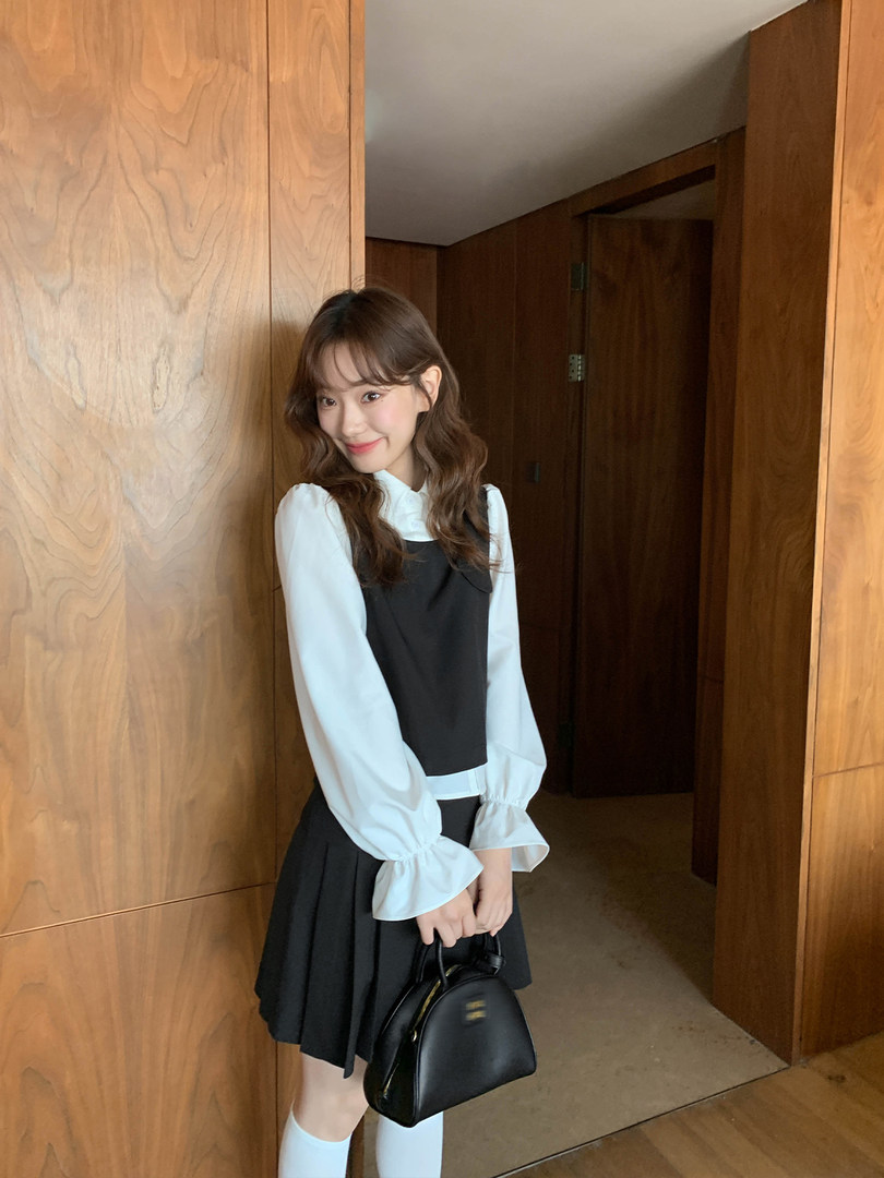 Woman wearing a black pinafore dress over a white blouse and holding a black handbag