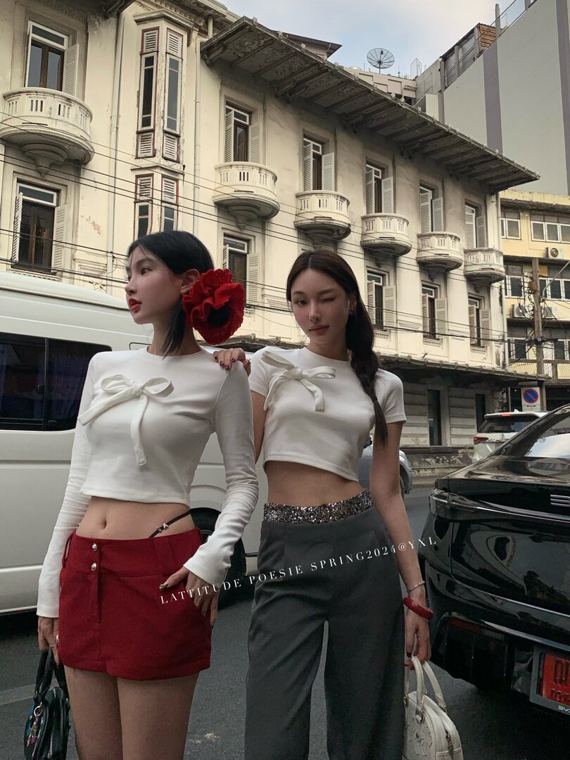 Two women wearing white bow-front crop tops styled with a red mini skirt and gray pants on a city street