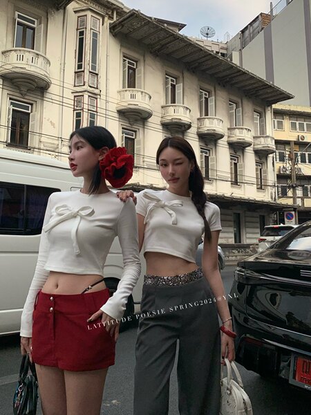 Two women wearing white bow-front crop tops styled with a red mini skirt and gray pants on a city street