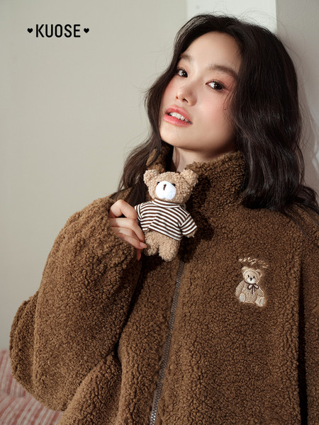Brown teddy fleece zip-up jacket worn by a model, with a bear patch and plush toy