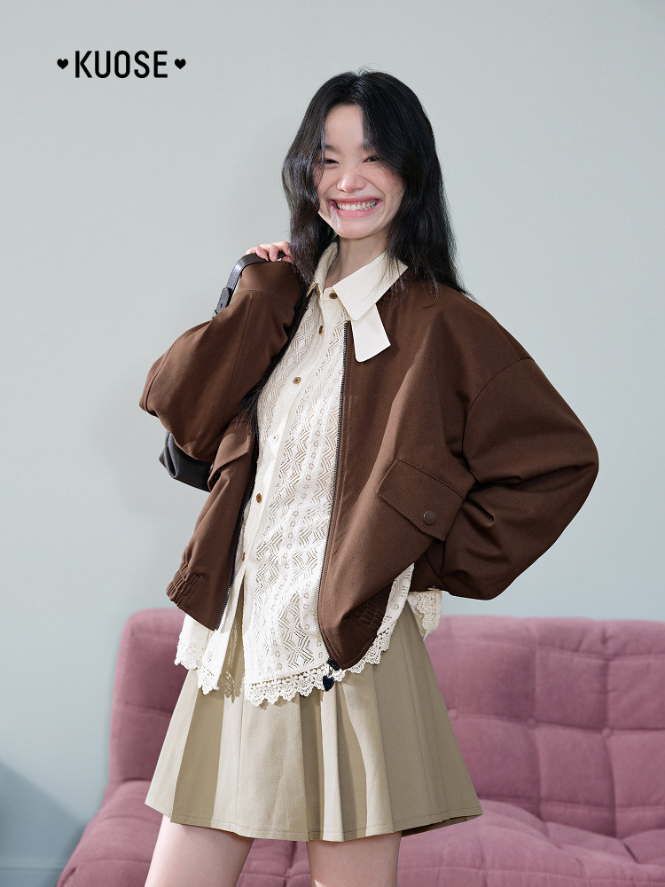 Model wearing a brown jacket over a white blouse and beige pleated skirt