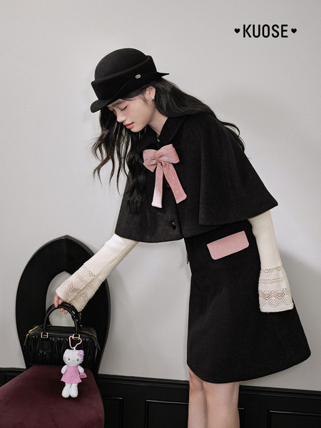 Woman modeling a black cape-style coat and skirt set with pink bow accents and a black hat