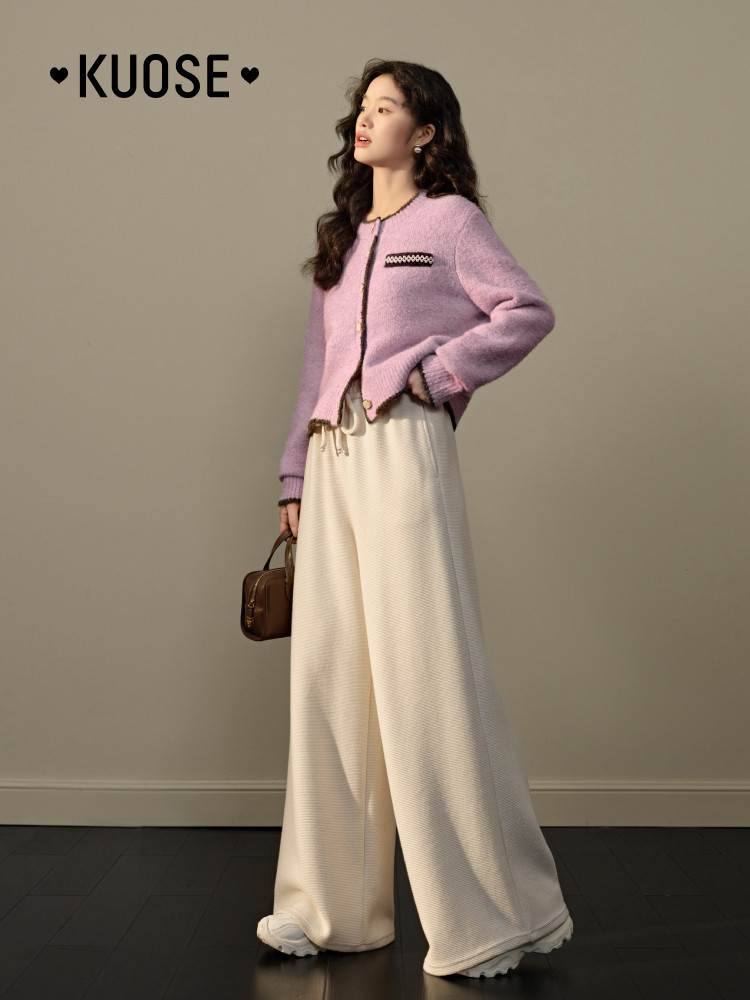 Model wearing a lavender cardigan with cream wide-leg pants and holding a small brown handbag