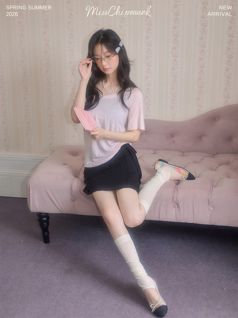 Model wearing a pink short-sleeve top, black skirt, and pastel flats in a bedroom setting