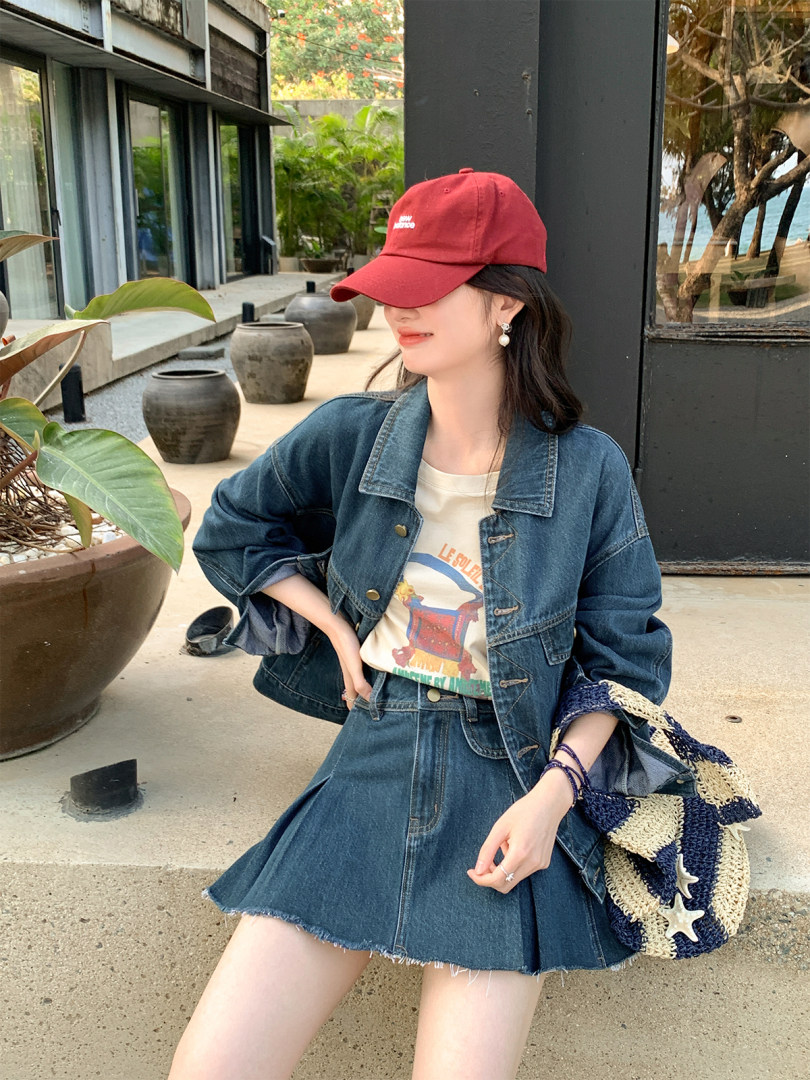 Woman wearing a denim jacket and pleated denim skirt with a red cap and woven beach bag