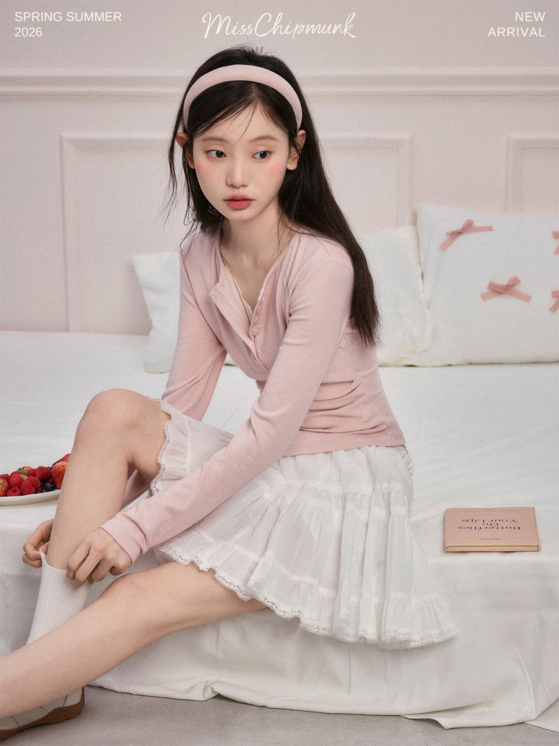 Young woman wearing a pink long-sleeve top and white ruffled skirt lounging on a bed