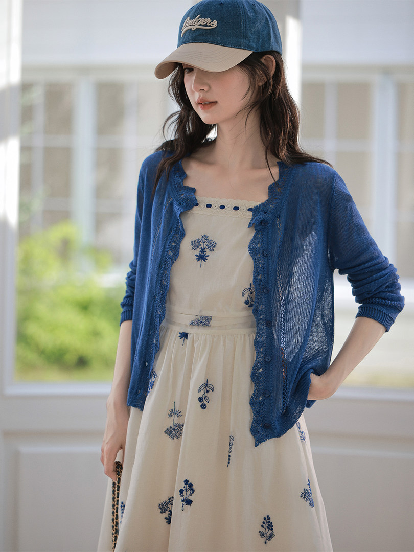Model wearing a blue cardigan over a cream embroidered dress with a baseball cap