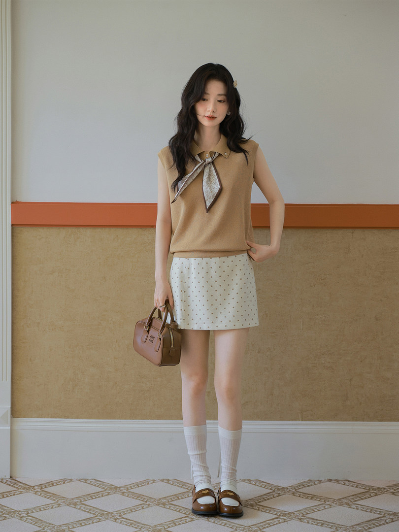 Model wearing a beige sleeveless knit top with a scarf tie, white polka dot mini skirt, and brown handbag