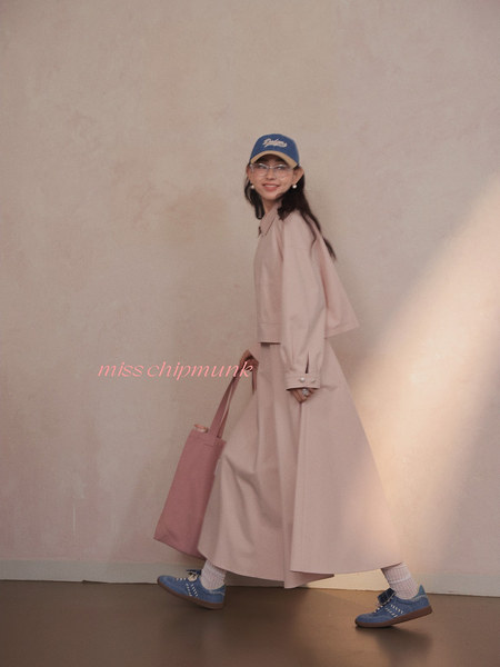 Woman wearing a pale pink long coat or dress with a blue cap, blue sneakers, and a pink tote bag