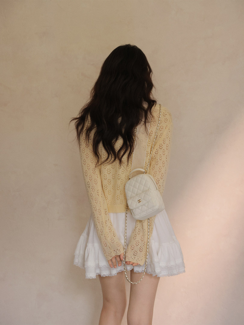 Model wearing a cream lace knit top with a white skirt and small quilted shoulder bag