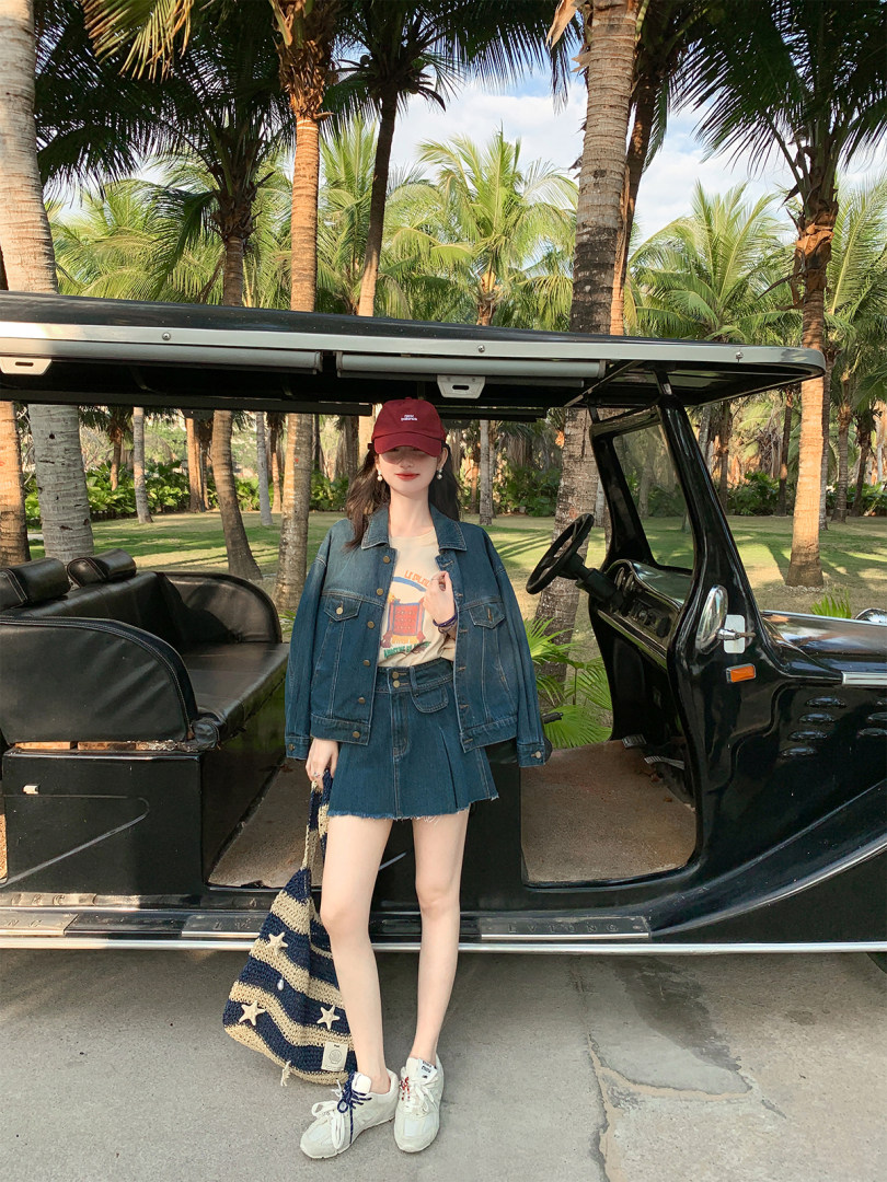 Woman wearing a denim jacket and skirt outfit with sneakers and a cap in a tropical outdoor setting