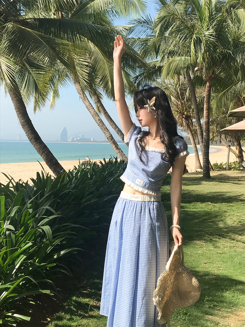 Model wearing a blue gingham two-piece outfit at the beach with a woven bag