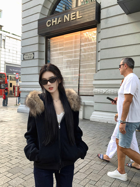 Woman wearing black zip-up jacket with fur-lined hood, standing on street in front of Chanel store
