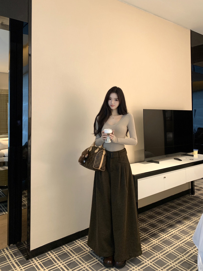 Woman wearing beige ribbed long-sleeve top and dark wide-leg pants, holding coffee cup and brown patterned handbag.