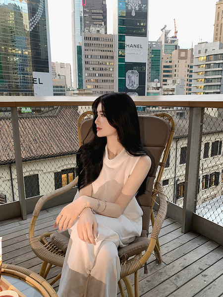 Woman wearing a white sleeveless top and pants, seated on a wicker chair on a balcony with a cityscape background.