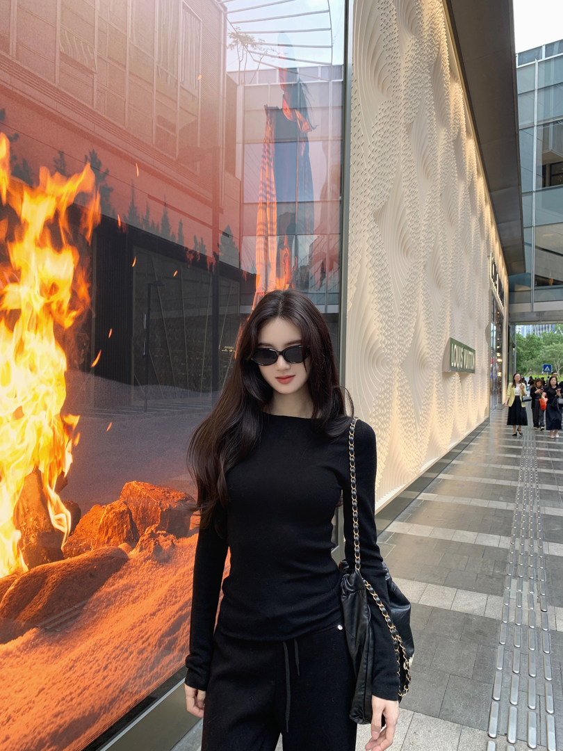 Woman wearing black long-sleeve top and black pants, carrying black chain-strap bag, standing outdoors near building with fire mural