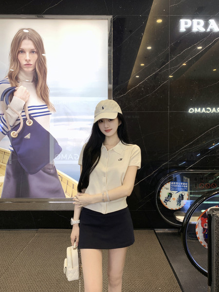 Woman wearing a beige polo shirt and black skirt, standing in front of a Prada advertisement.