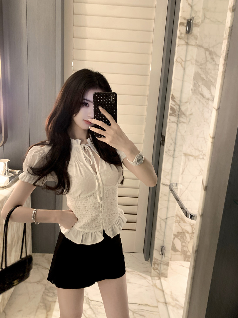 Woman taking mirror selfie wearing white blouse and black shorts