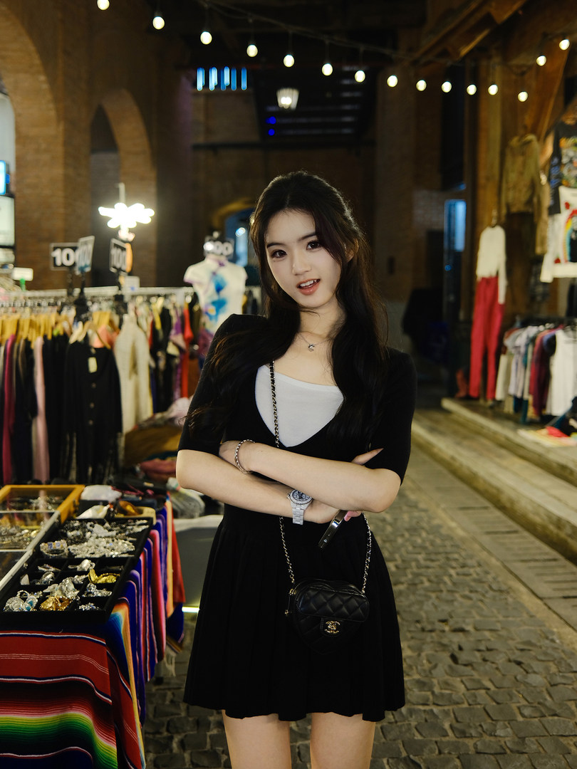 Woman wearing a black dress with a white top underneath, standing in a market setting.