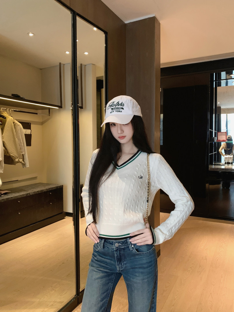 Woman wearing white cable knit sweater, blue jeans, and a white cap with text.