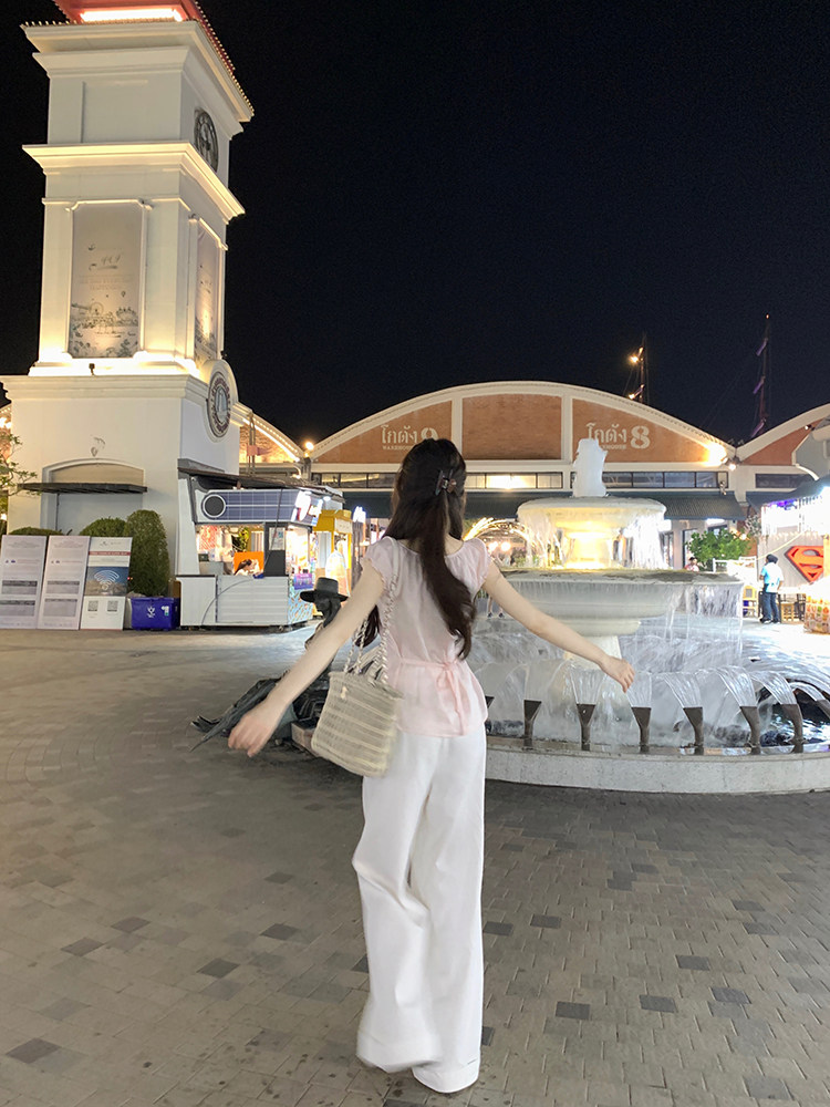 Woman wearing pink top and white pants standing in front of fountain at night
