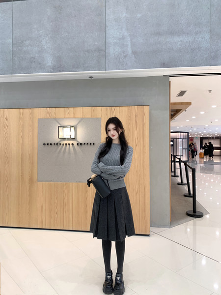 Woman wearing gray cable knit sweater, black pleated skirt, black tights, and black platform loafers in a mall setting
