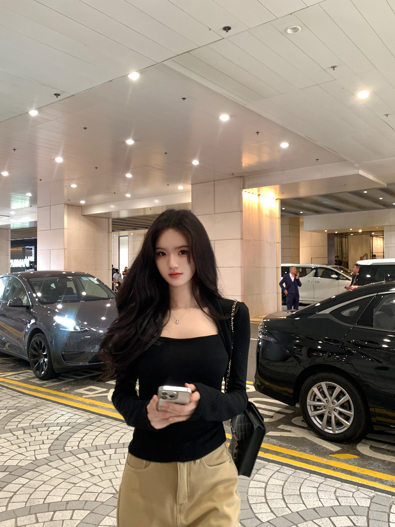 Woman wearing black long-sleeve top and beige pants, holding phone, standing in front of cars and building entrance