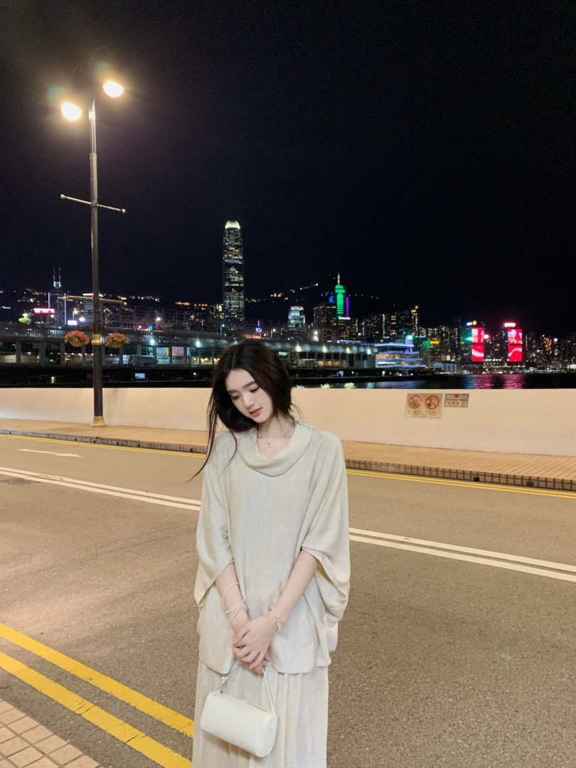 Woman wearing a light-colored loose-fitting dress with cowl neckline, standing on a city street at night with skyline background