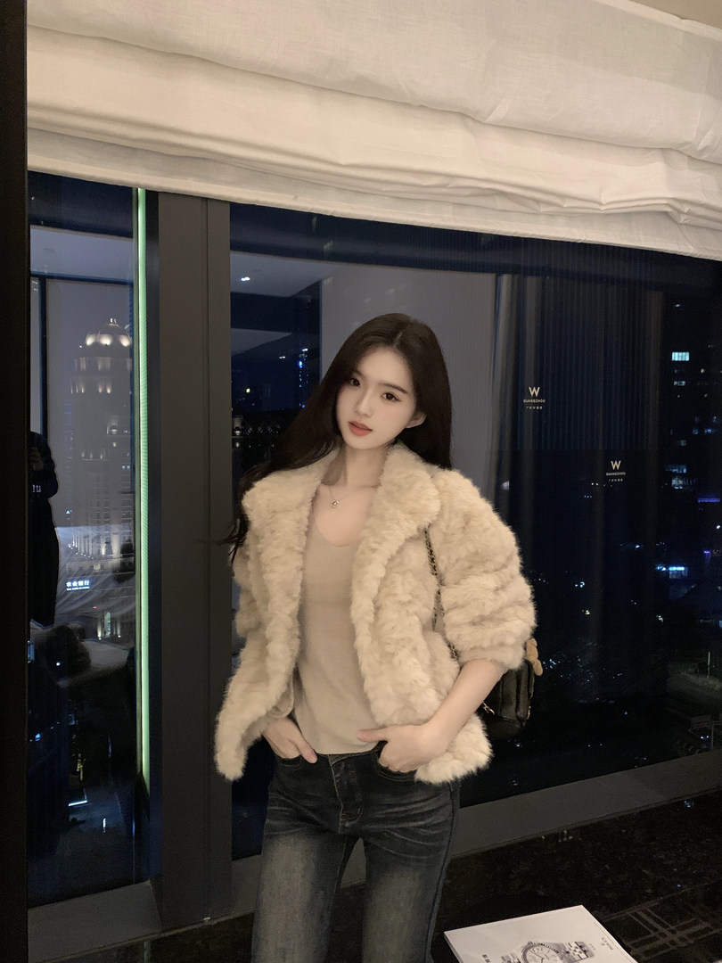 Woman wearing a beige faux fur jacket with jeans, standing indoors near a window with city lights in the background.