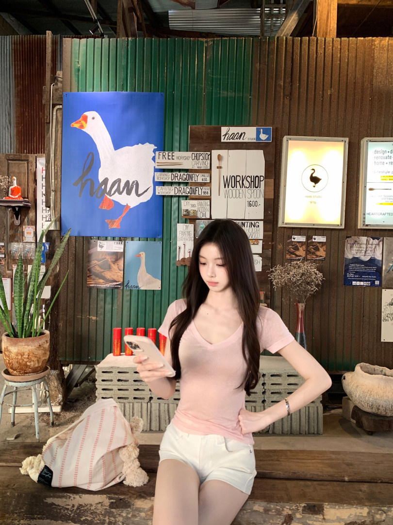 Woman wearing a pink V-neck top and white shorts, posing with a phone in a rustic workshop setting.