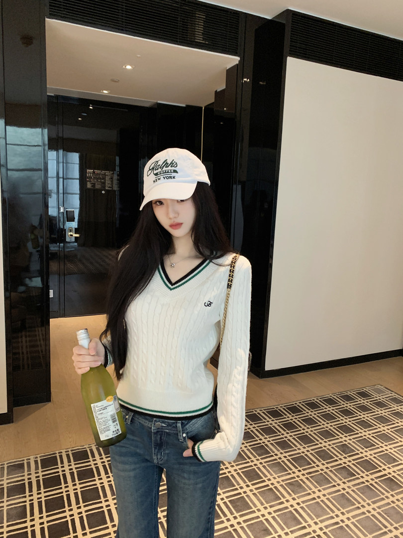 Woman wearing white cable knit sweater with green and black trim, blue jeans, and white cap, holding a bottle.