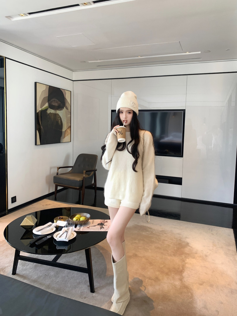 Model wearing cream knit sweater, matching shorts, beanie, and knee-high boots in a modern living room