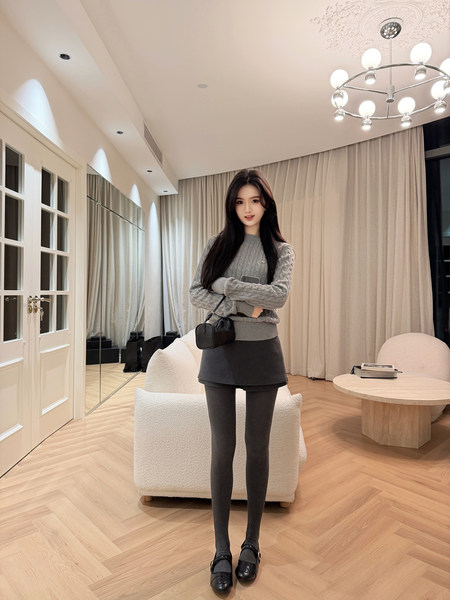 Woman wearing a gray cable knit sweater, black skirt, and tights, standing in a modern living room.