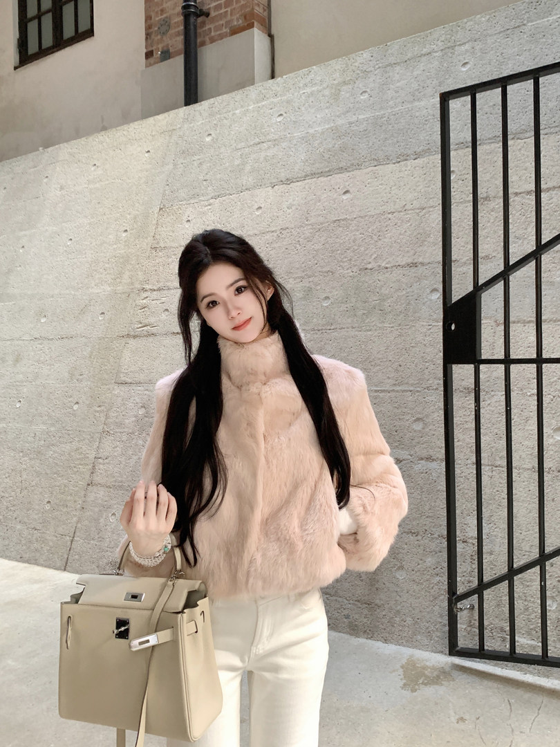 Woman wearing a light pink faux fur coat with white pants and holding a beige handbag, standing against a concrete wall.