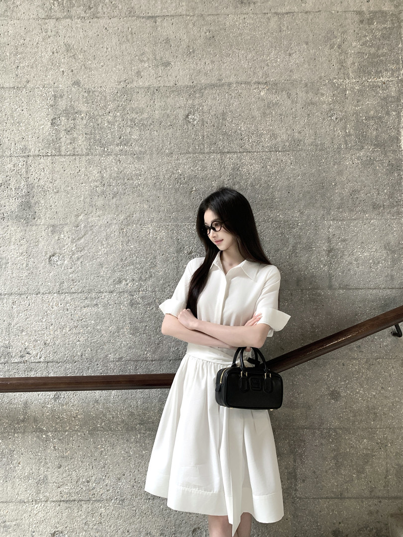 Woman wearing a white shirt dress with rolled sleeves, standing against a concrete wall with a black handbag.