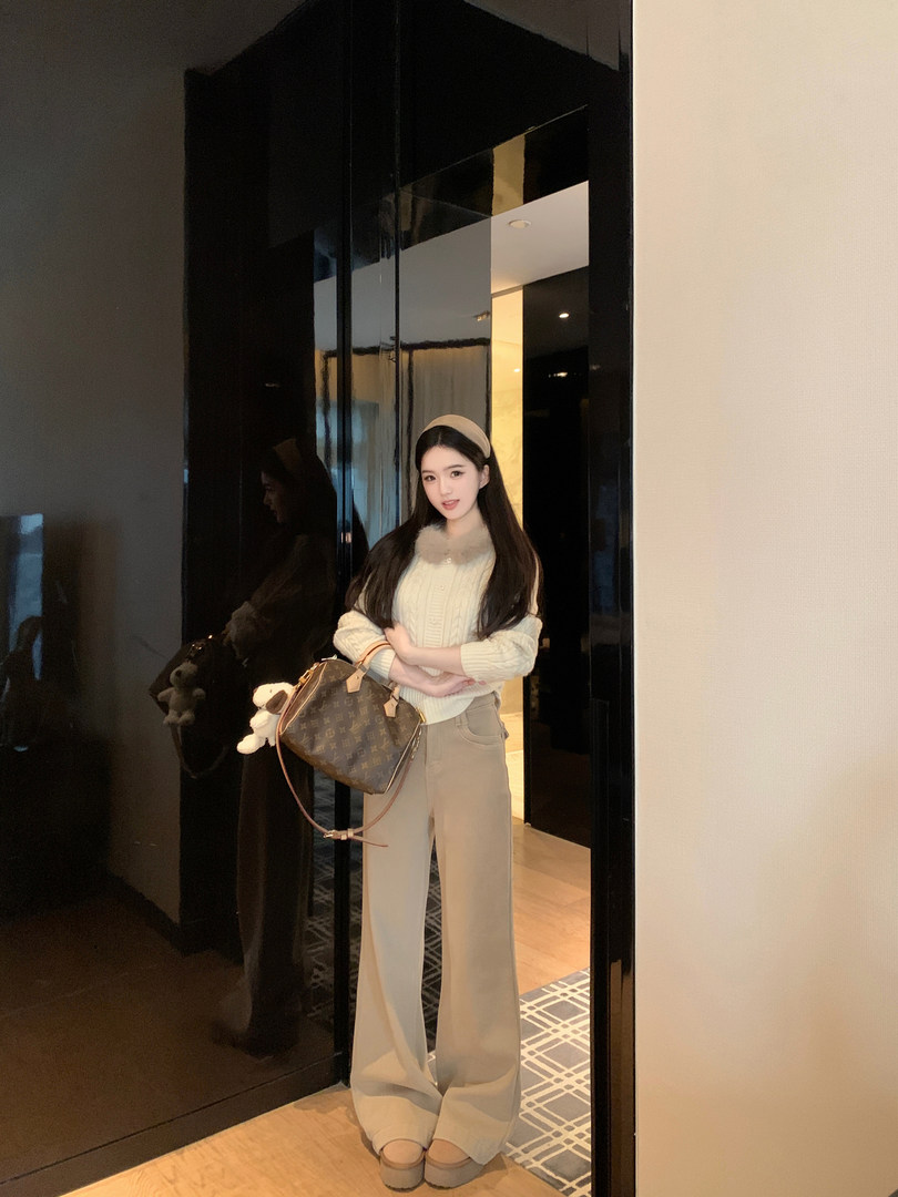 Woman wearing cream cable knit sweater with faux fur collar, beige wide-leg pants, and carrying a patterned handbag, standing in front of a mirror.