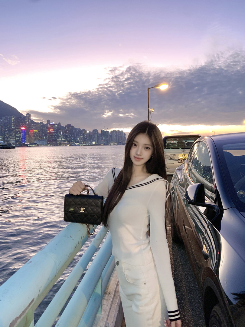 Woman holding a black quilted handbag while standing by a waterfront with a city skyline in the background.