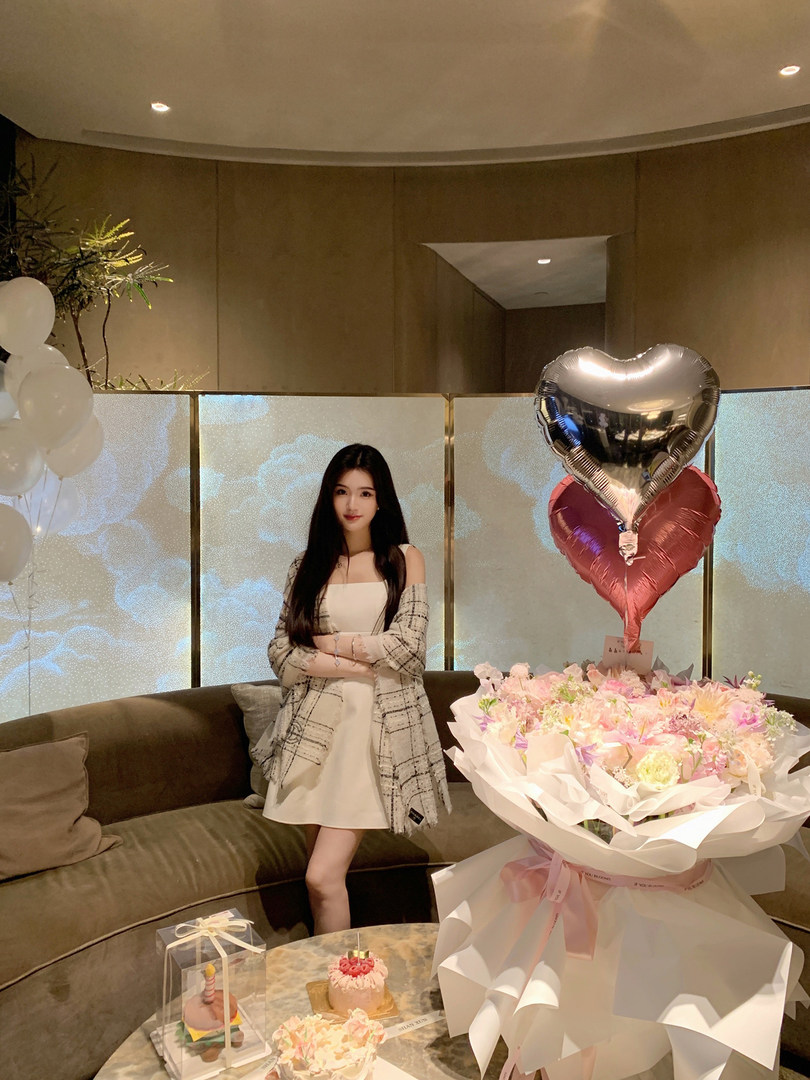 Woman wearing a white dress with a plaid cardigan, standing next to a bouquet of flowers and heart-shaped balloons in a decorated room.