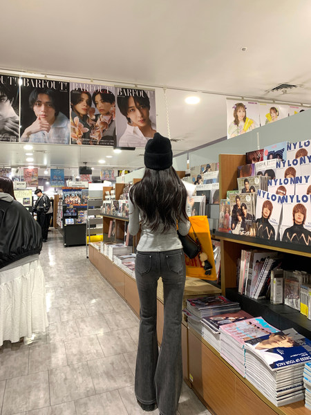 Woman wearing gray top and dark flared jeans from behind in a magazine store aisle