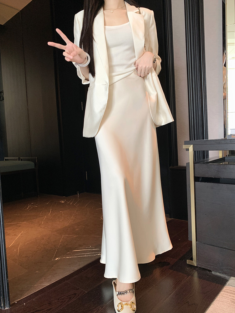 Woman wearing a cream satin skirt and matching blazer with white heels, posing indoors