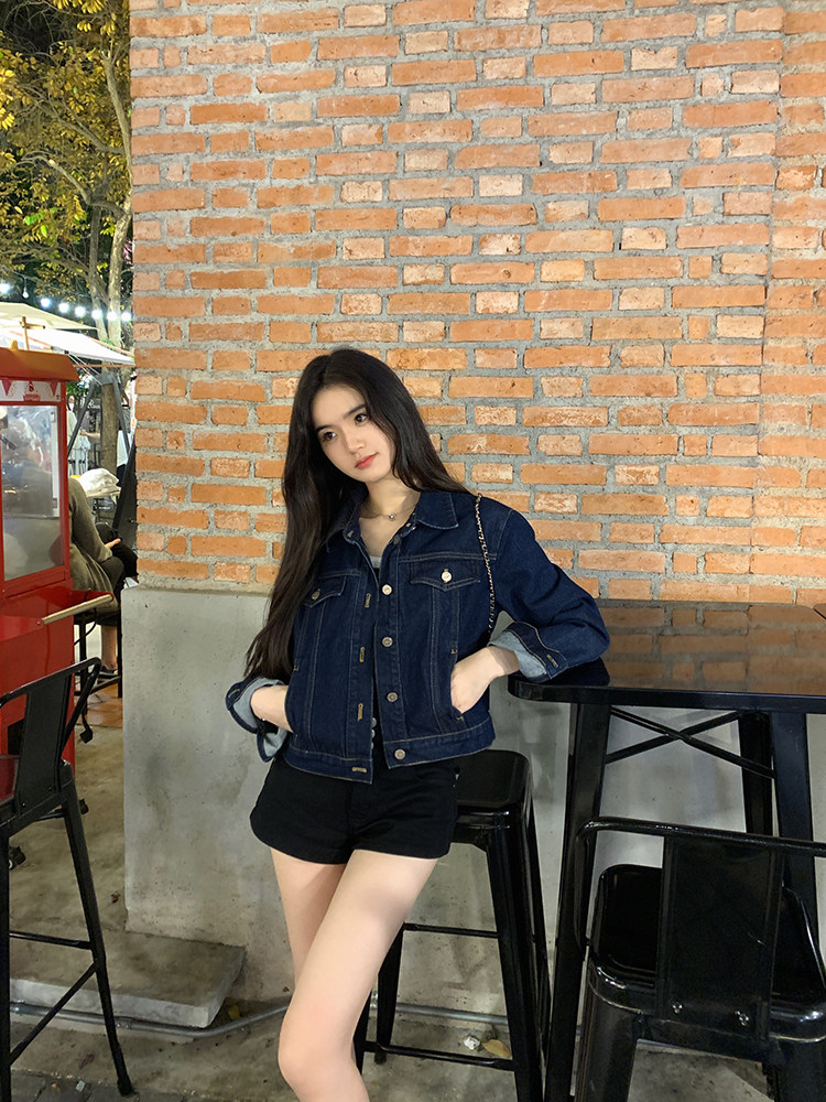 Woman wearing denim jacket and black shorts leaning against a brick wall