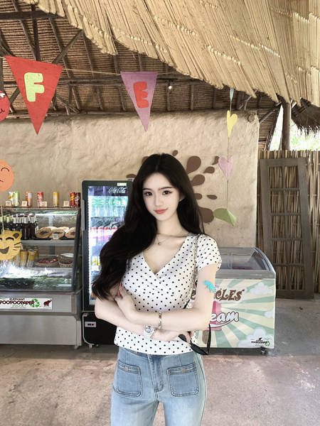 Woman wearing a white polka dot top and light blue jeans standing in front of a rustic shop with decorations.