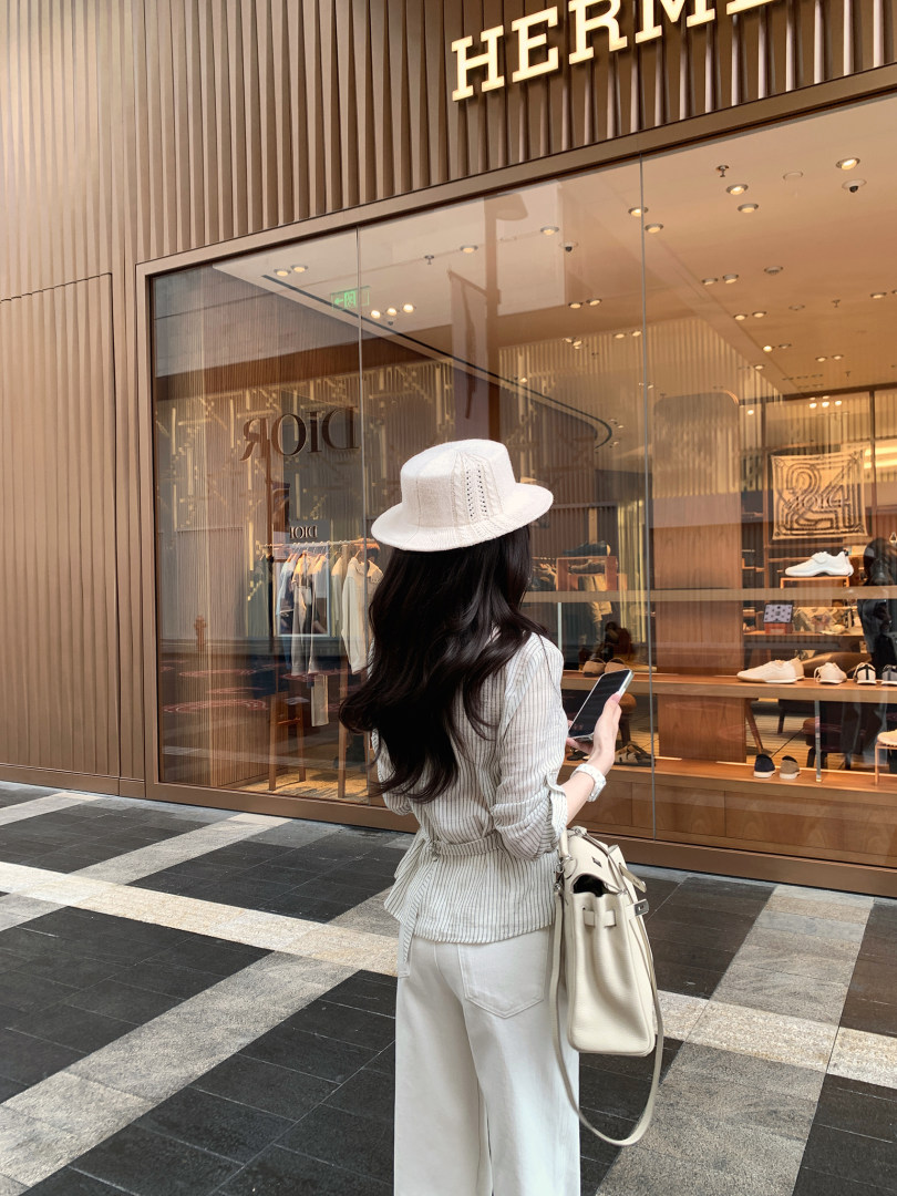 Woman wearing a white hat, striped shirt, and white pants, carrying a beige handbag, standing in front of a luxury store.