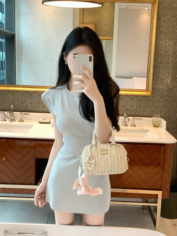 Woman taking mirror selfie wearing light blue knit dress and holding cream handbag with plush keychain