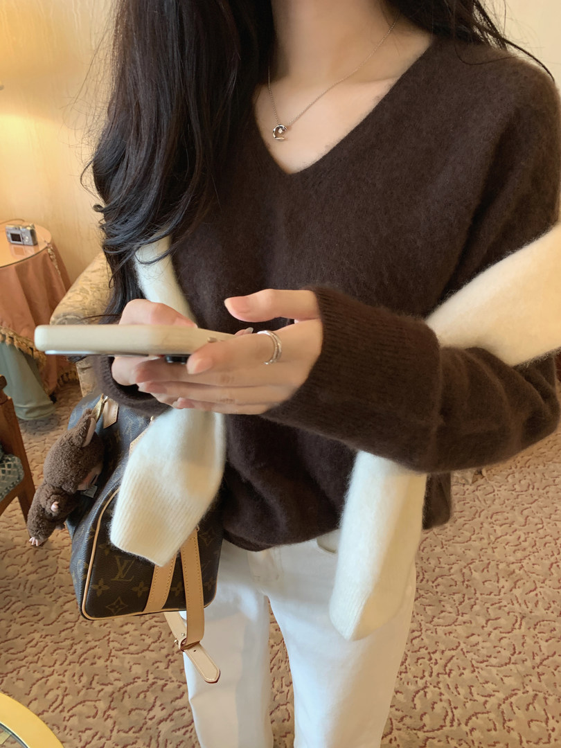Woman wearing a brown V-neck sweater with white pants and holding a phone