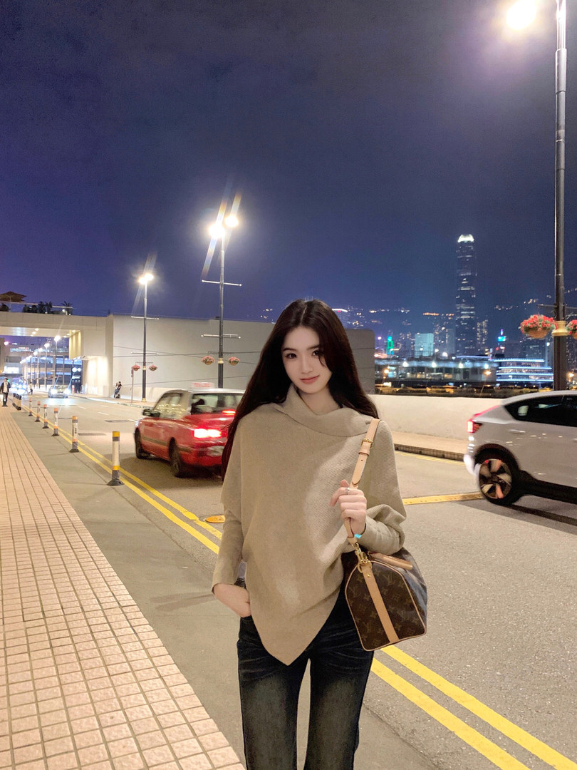 Woman wearing a beige sweater and jeans, holding a designer bag on a city street at night.