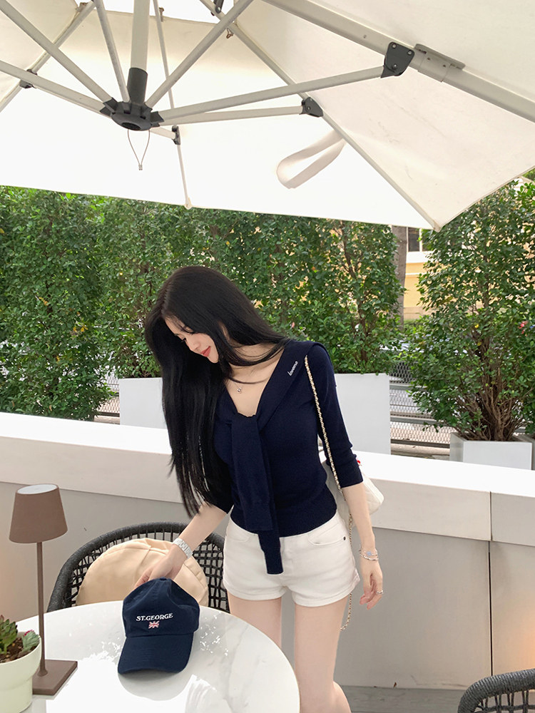 Woman wearing navy blue wrap top and white shorts, holding a navy cap with 'ST.GEORGE' text, under an outdoor umbrella.