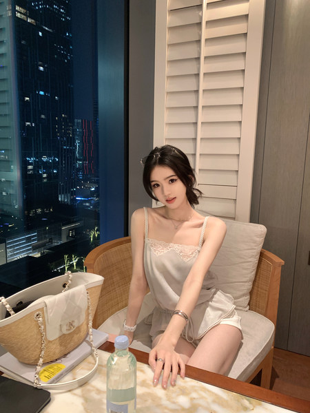 Woman wearing a light-colored lace-trimmed camisole and shorts set, seated indoors with city skyline view at night