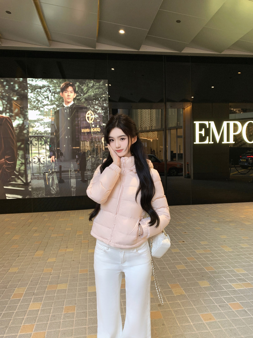 Woman wearing a light pink puffer jacket and white pants, posing outdoors in front of a store.