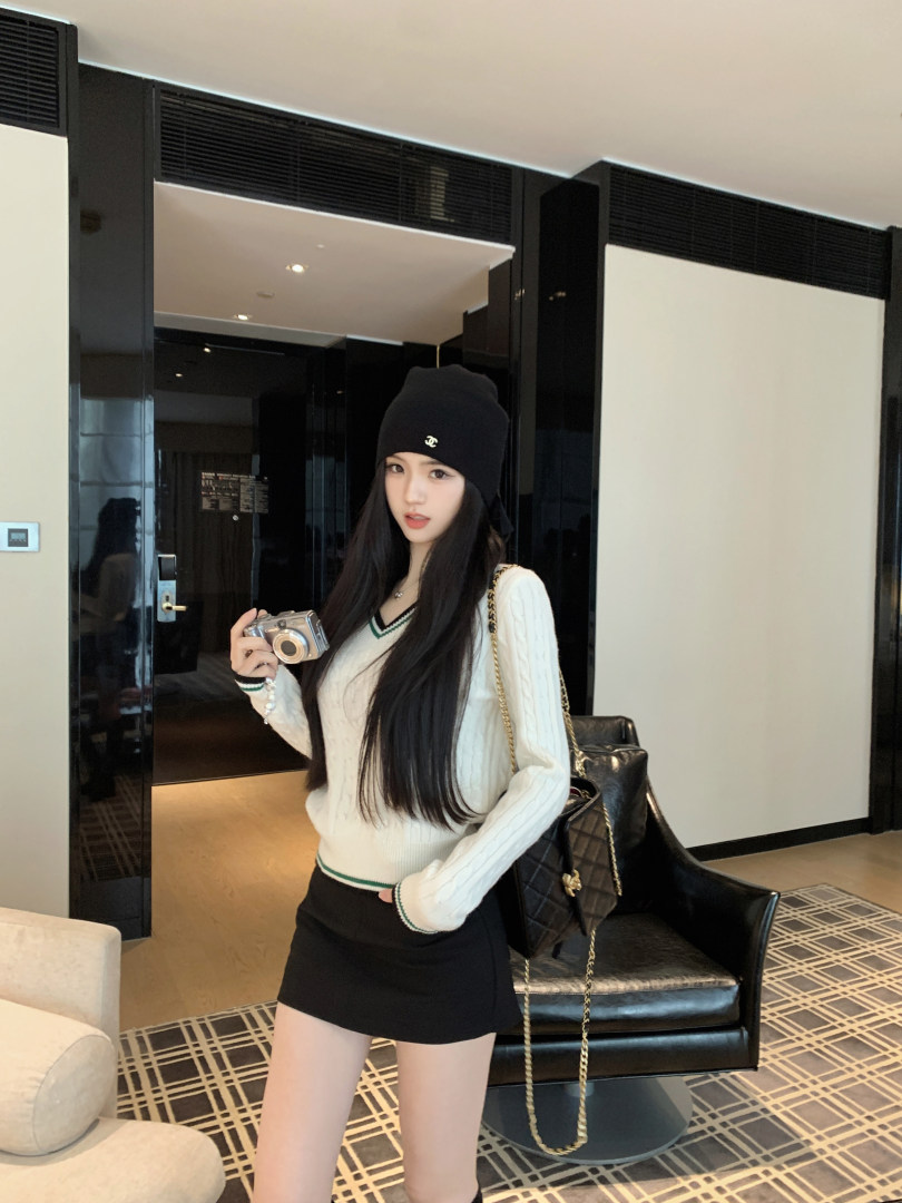Woman wearing white cable knit sweater with green trim, black mini skirt, and black beanie with logo, holding camera in hotel room